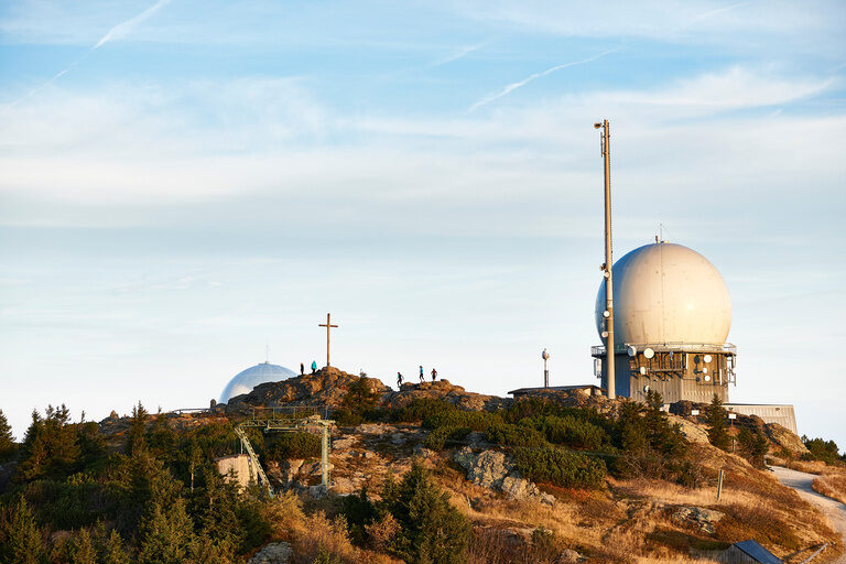Gipfel des Großen Arber im Bayerischen Wald mit Radaranlagen, Wanderern und Kreuz bei warmem Licht und klarem Himmel.